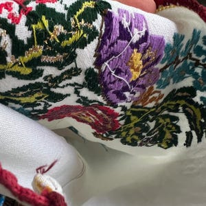 May include: Close-up of white fabric with vibrant floral embroidery. The embroidery includes purple, red, green, and yellow flowers and foliage. A red and gold crocheted border trims the edge. The fabric is held in a hand.