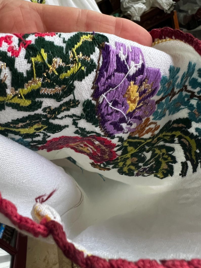 May include: Close-up of white fabric with vibrant floral embroidery. The embroidery includes purple, red, green, and yellow flowers and foliage. A red and gold crocheted border trims the edge. The fabric is held in a hand.