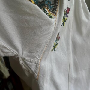 May include: White embroidered shirt with colourful floral and geometric patterns. The neckline and sleeves feature detailed stitching in various colours, including green, yellow, and red. The shirt's fabric appears to be a soft, lightweight material.