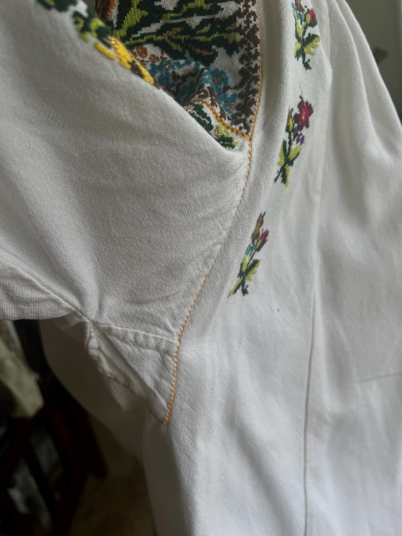May include: White embroidered shirt with colourful floral and geometric patterns. The neckline and sleeves feature detailed stitching in various colours, including green, yellow, and red. The shirt's fabric appears to be a soft, lightweight material.