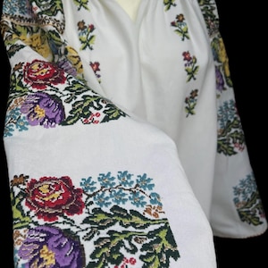 May include: White embroidered blouse with colourful floral patterns on the sleeves and bodice. The embroidery features red, purple, blue, and green flowers and leaves. The neckline is trimmed with orange and the sleeves have a flared design.