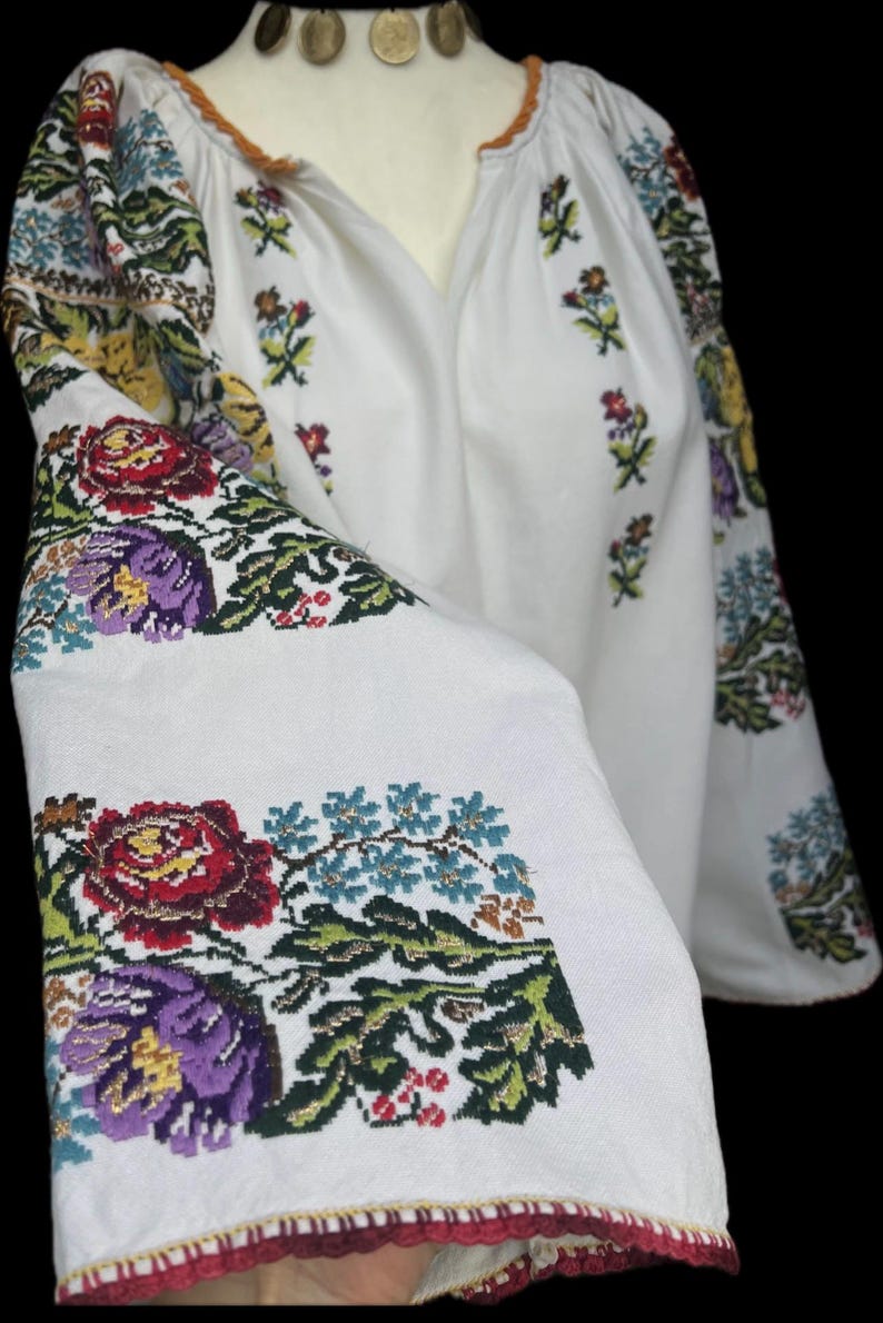 May include: White embroidered blouse with colourful floral patterns on the sleeves and bodice. The embroidery features red, purple, blue, and green flowers and leaves. The neckline is trimmed with orange and the sleeves have a flared design.