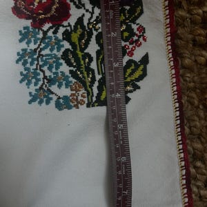 May include: White fabric with embroidered floral design in red, purple, blue, and gold. The edge is trimmed with red and gold stitching. A ruler shows the fabric is approximately 53 cm long.