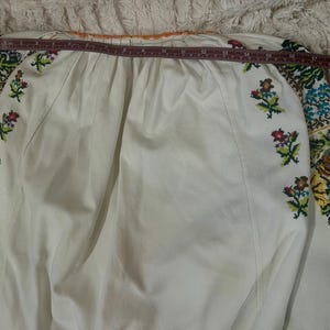 May include: White long-sleeved blouse with colourful floral embroidery on the sleeves and small floral accents on the front. The blouse has a round neckline and is measured with a tape measure. The embroidery features shades of blue, green, yellow, and pink.