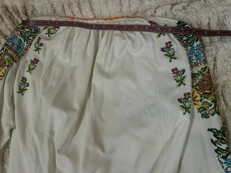 May include: White long-sleeved blouse with colourful floral embroidery on the sleeves and small floral accents on the front. The blouse has a round neckline and is measured with a tape measure. The embroidery features shades of blue, green, yellow, and pink.