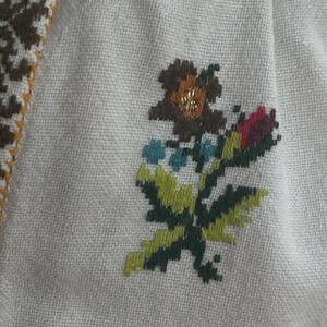 May include: Close-up of cream fabric with cross-stitch embroidery. The embroidery depicts a floral design in brown, green, blue, and red. A border with a geometric pattern in brown, yellow, and white is visible.