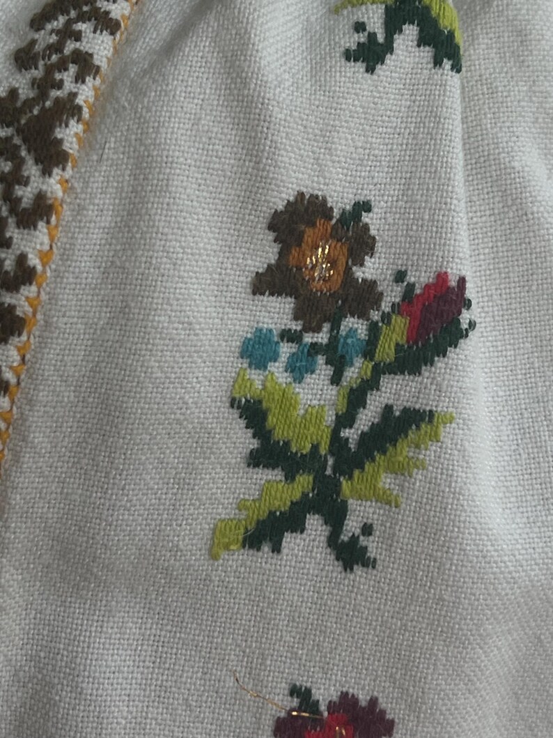 May include: Close-up of cream fabric with cross-stitch embroidery. The embroidery depicts a floral design in brown, green, blue, and red. A border with a geometric pattern in brown, yellow, and white is visible.