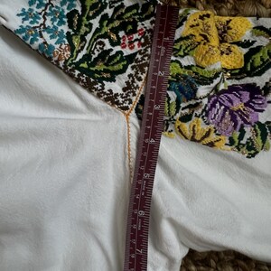 May include: White embroidered blouse with colourful floral patterns on the sleeves. The embroidery includes blue, yellow, purple, and green flowers with gold accents. A ruler shows a measurement of approximately 20 cm.