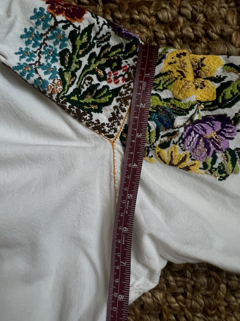 May include: White embroidered blouse with colourful floral patterns on the sleeves. The embroidery includes blue, yellow, purple, and green flowers with gold accents. A ruler shows a measurement of approximately 20 cm.