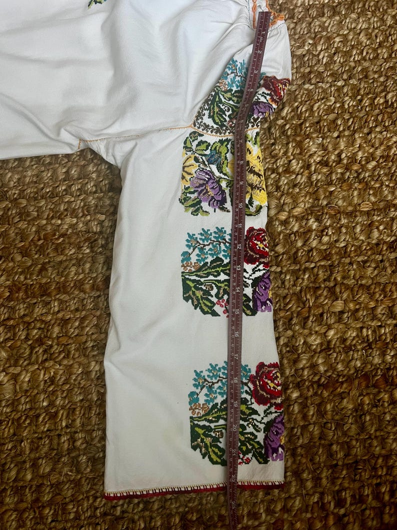 May include: White embroidered sleeve with colourful floral patterns in red, purple, yellow, and blue. The sleeve is measured with a ruler, showing a length of approximately 50 cm. The embroidery is detailed and vibrant.