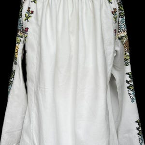 May include: White long-sleeved blouse with floral embroidery on the sleeves. The embroidery features red, green, blue, and yellow flowers and leaves. The blouse has a gathered neckline and flared sleeves, with red trim at the cuffs.