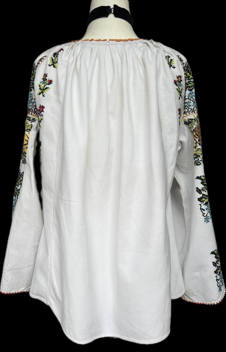 May include: White long-sleeved blouse with floral embroidery on the sleeves. The embroidery features red, green, blue, and yellow flowers and leaves. The blouse has a gathered neckline and flared sleeves, with red trim at the cuffs.