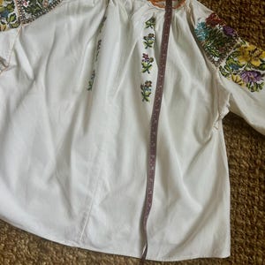 May include: White embroidered blouse with colourful floral patterns on the sleeves and bodice. The blouse has long sleeves and a round neckline with orange trim. A measuring tape is placed on the blouse, showing its length.