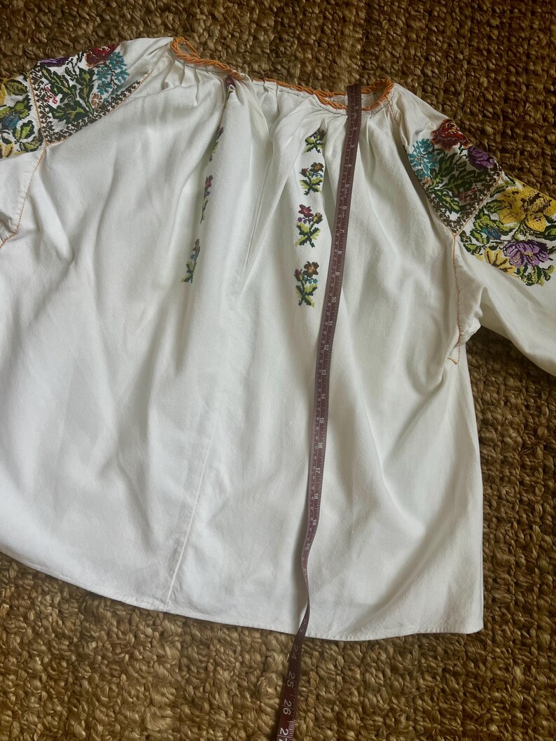 May include: White embroidered blouse with colourful floral patterns on the sleeves and bodice. The blouse has long sleeves and a round neckline with orange trim. A measuring tape is placed on the blouse, showing its length.