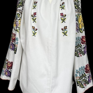 May include: White embroidered blouse with long sleeves. The sleeves and upper chest feature colourful floral embroidery in red, yellow, purple, and green. The neckline is a V-shape, and the sleeves have a red trim. The blouse is made of a soft, white fabric.
