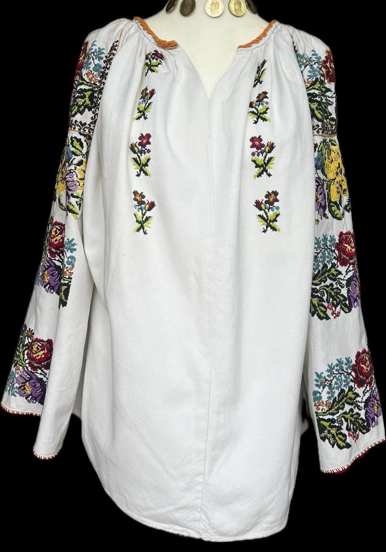 May include: White embroidered blouse with long sleeves. The sleeves and upper chest feature colourful floral embroidery in red, yellow, purple, and green. The neckline is a V-shape, and the sleeves have a red trim. The blouse is made of a soft, white fabric.