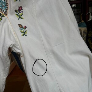 May include: Close-up of a white embroidered blouse. The blouse has colourful floral embroidery on the collar and sleeves. The sleeves are long and the fabric appears to be a lightweight cotton or linen.
