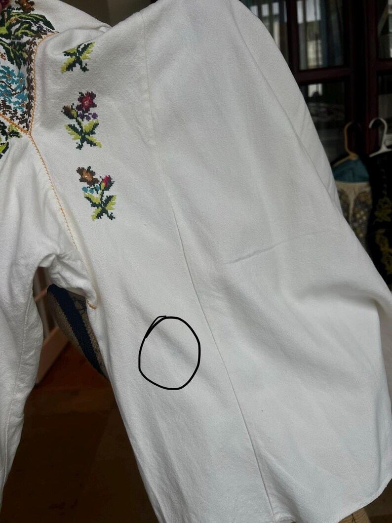 May include: Close-up of a white embroidered blouse. The blouse has colourful floral embroidery on the collar and sleeves. The sleeves are long and the fabric appears to be a lightweight cotton or linen.