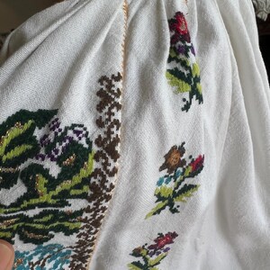 May include: White embroidered blouse with colourful floral patterns. The embroidery features green, red, purple, and gold accents. The blouse has a gathered neckline and detailed stitching along the seams, showcasing traditional craftsmanship.