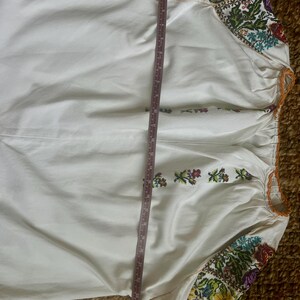 May include: White long-sleeved blouse with colourful floral embroidery on the sleeves and neckline. The blouse has a gathered neckline and a measurement tape is placed vertically across the garment. The embroidery features shades of purple, yellow, green, and red.