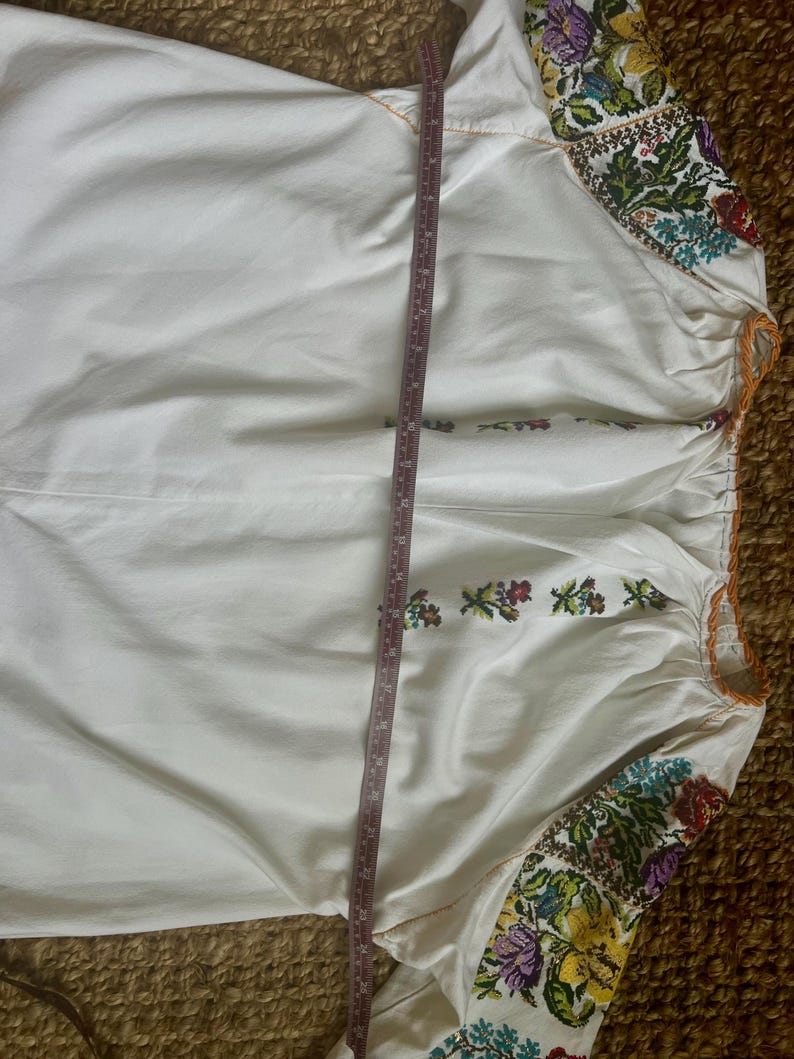 May include: White long-sleeved blouse with colourful floral embroidery on the sleeves and neckline. The blouse has a gathered neckline and a measurement tape is placed vertically across the garment. The embroidery features shades of purple, yellow, green, and red.
