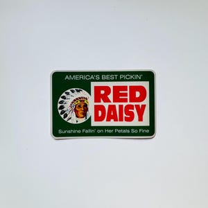May include: A rectangular sticker with a dark green border and white background. It features a circular illustration of a person in a feathered headdress, the red text RED DAISY, and the phrases AMERICA'S BEST PICKIN' and Sunshine Fallin' on Her Petals So Fine.