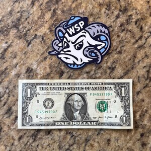 May include: A die-cut sticker featuring a stylized ram head mascot with large curved horns, wearing a small blue hat, and displaying the letters WSP in white. The sticker is positioned above a standard United States one-dollar bill for scale.