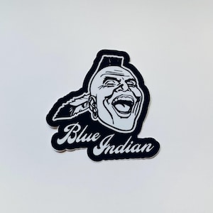 May include: A black and white sticker featuring a stylized illustration of a person with a mohawk hairstyle and a feather behind the ear. The person has an open-mouthed expression. Below the illustration, the text Blue Indian is written in a white script font.
