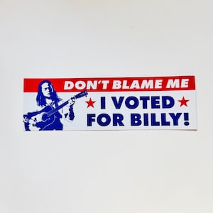 May include: A rectangular bumper sticker with a red top stripe and white background. It features a blue silhouette of a person playing an acoustic guitar. The text reads: DON'T BLAME ME, I VOTED FOR BILLY!, with a red star on each side of the text.