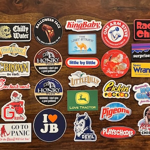 May include: A collection of colorful stickers with various designs and text, including "Chilly Water," "KingBaby," "Radio Child," and "Tie Your Shoes." The stickers are arranged on a wooden surface, showcasing a variety of themes and styles.