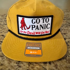 May include: A mustard yellow baseball cap with a black rope trim across the brim. A rectangular patch features a red figure, black text reading GO TO PANIC, and red text reading Or the Devil Will Get You!. A Richardson R-ACTIVE UPF 50+ sticker is attached.