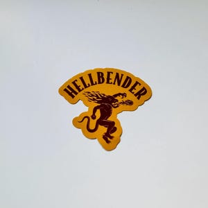 May include: A die-cut sticker with a mustard yellow background featuring a dark brown graphic of a creature with a long tail and flames on its head, breathing fire. The text HELLBENDER is printed in dark brown capital letters above the creature.
