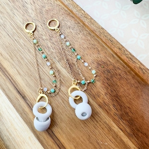 May include: A pair of long, gold chain earrings with white and green beads. The earrings have a white, round charm at the bottom.