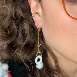 May include: Gold dangle earrings with a white ceramic charm. The charm is a series of interlocking circles. The earrings also feature a small gold star charm.