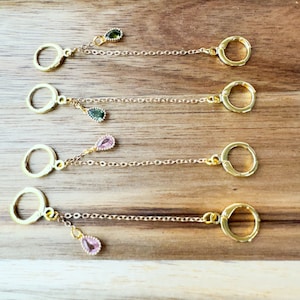 May include: Five gold hoop earrings with dangling chains and small gemstone accents. The gemstones are green and pink.