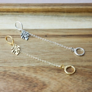 以下が含まれることがあります： ゴールドとシルバーの2つのドロップピアス。それぞれに小さなモンステラの葉のチャームが付いています。ピアスはチェーンから吊り下げられ、小さなフープで終わります。ピアスは木製の表面に展示されています。