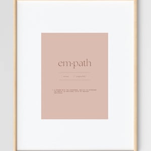 May include: A framed print featuring the word "em.path" in a stylish serif font, set against a muted rose-colored background. The definition of "empath" is included below the word. The frame is a light wood color.
