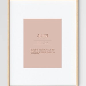 May include: A printable art print with a light pink background and a brown frame. The print features the word "aura" in a brown font with a definition below. The definition is in a smaller font size and explains the meaning of the word "aura".