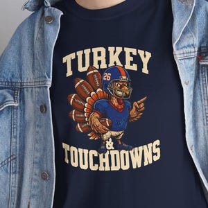 May include: A dark navy blue t-shirt featuring a graphic of a turkey wearing a football helmet and jersey, holding a football. The text reads TURKEY & TOUCHDOWNS in a cream-coloured font. The shirt is worn under a light blue denim jacket.