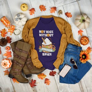 May include: A purple t-shirt with the text "Not Nice Without Pumpkin Spice" and a cartoon pumpkin spice latte. The shirt is laid out on a white wood background with fall-themed decorations, including pumpkins, leaves, and boots.