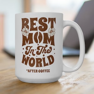 Op de afbeelding: Witte keramische koffiemok met een bruine afbeelding met de tekst "Best Mom In The World *After Coffee" met twee witte bloemen aan weerszijden van het woord "Mom".