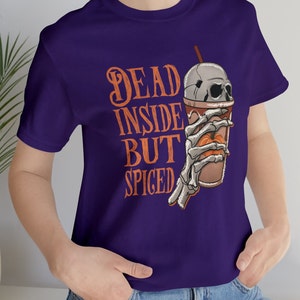 May include: Purple t-shirt featuring a skeleton hand holding a pumpkin spice latte. The text on the shirt reads "Dead Inside But Spiced".
