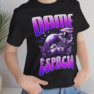 May include: A black t-shirt with a purple and pink gradient design featuring the text "DAME ESPACIO" and an astronaut sitting on a moon.