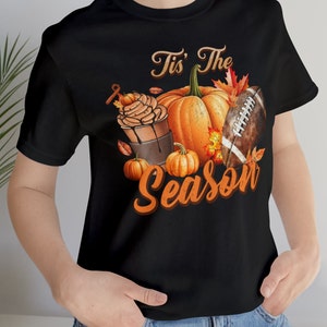 Pode incluir: Camiseta preta com um design gráfico que apresenta uma abóbora, uma bola de futebol americano e uma bebida de café. O texto "Tis' The Season" está escrito em uma fonte de estilo vintage.