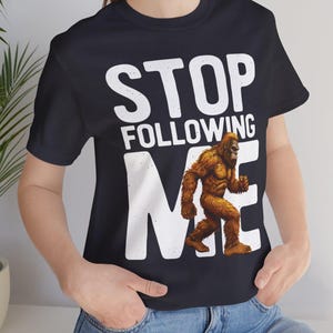 May include: Black t-shirt with the text "STOP FOLLOWING ME" in white block letters. A brown Bigfoot graphic is in the center. The shirt is a crew neck style.