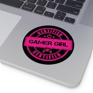 Peut inclure: Un autocollant rond avec un fond noir et un design rose. Le design comprend les mots "CERTIFIED GAMER GIRL" dans une police vieillie, ainsi qu'une icône de manette de jeu et des étoiles. L'autocollant est sur un ordinateur portable.