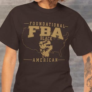 May include: A dark brown t-shirt with the text "FOUNDATIONAL FBA BLACK AMERICAN" in a gold color. The design includes a map of the United States with a fist graphic.