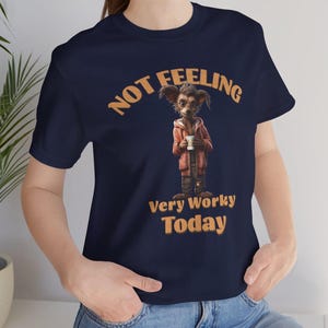 May include: A navy blue short-sleeve t-shirt featuring a graphic of a small, furry creature wearing a red hoodie and holding a white coffee cup. The text above and below the creature reads, "NOT FEELING Very Worky Today" in orange letters.