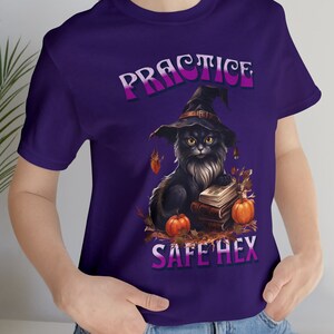 Puede incluir: Camiseta morada con un gato negro con un sombrero de bruja y sosteniendo libros. El texto "PRACTICE SAFE HEX" está impreso en la camiseta.