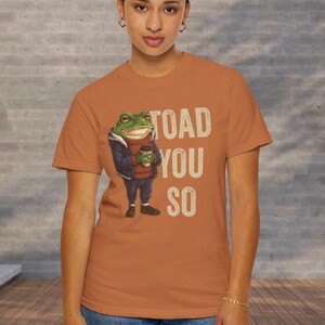May include: An orange t-shirt with a cartoon frog in a sweater holding a coffee cup. The text "TOAD YOU SO" is printed in cream. The shirt is worn by a person wearing blue jeans.